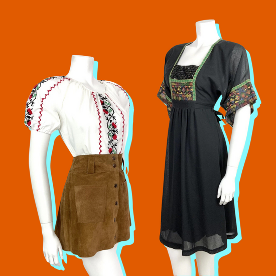 two mannequins wear vintage 1960s 1970s folk style clothing. The mannequin on the left wear a white puff sleeve blouse embroidered with red roses, and is wearing a brown suede mini skirt. The mannequin on the right wears a black short sleeve empire line smock dress embroidered with rainbow swirls and green trim